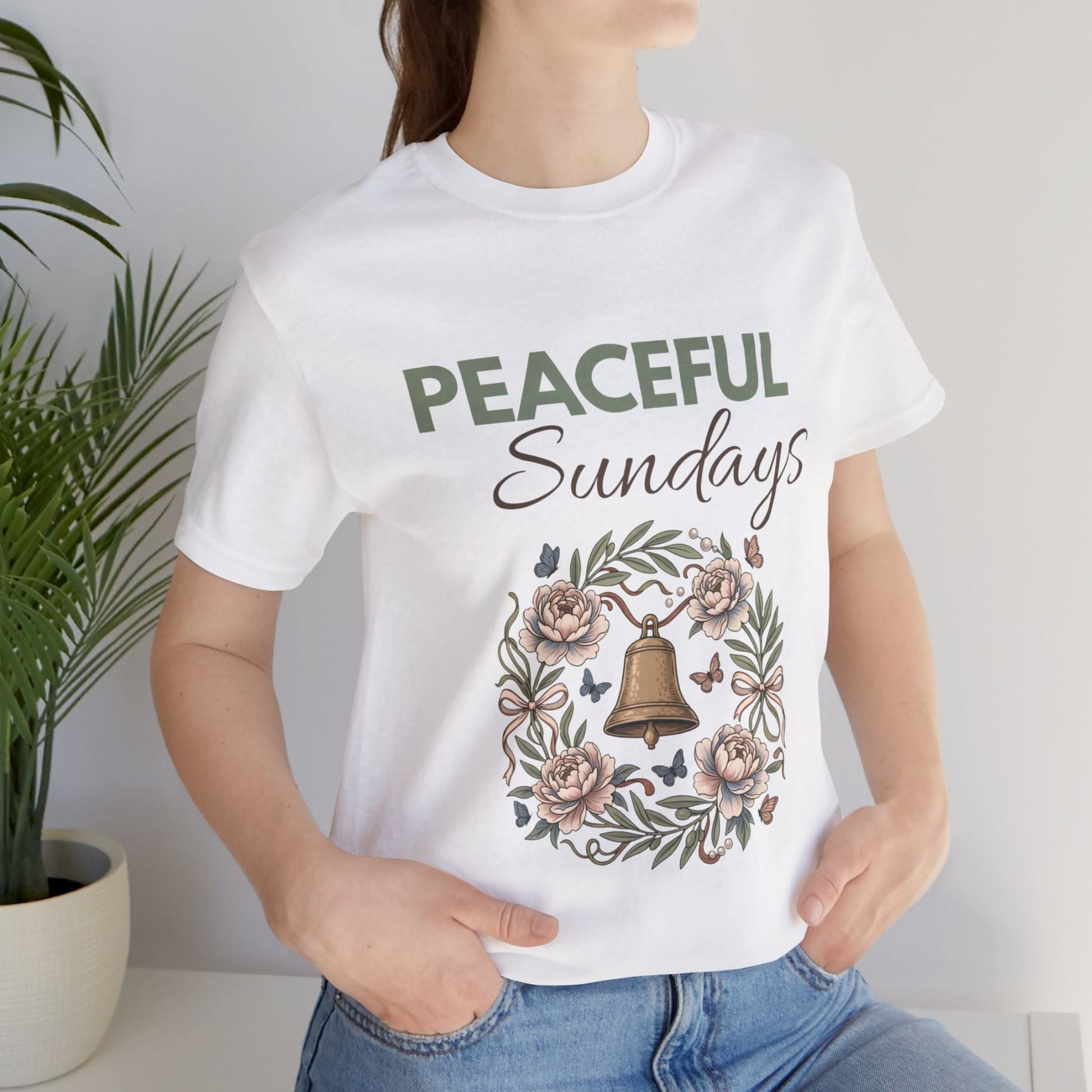Person wearing a white t-shirt with 'Peaceful Sundays' design, standing next to a plant.