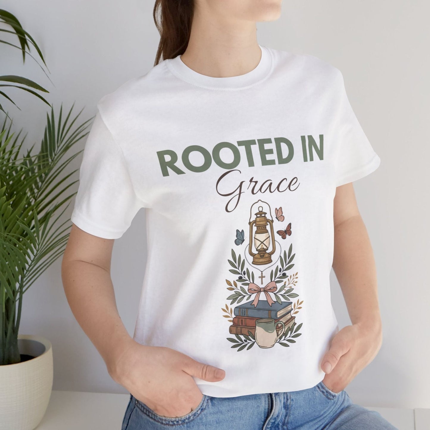 Person wearing a white t-shirt with 'Rooted in Grace' design, standing next to a plant.