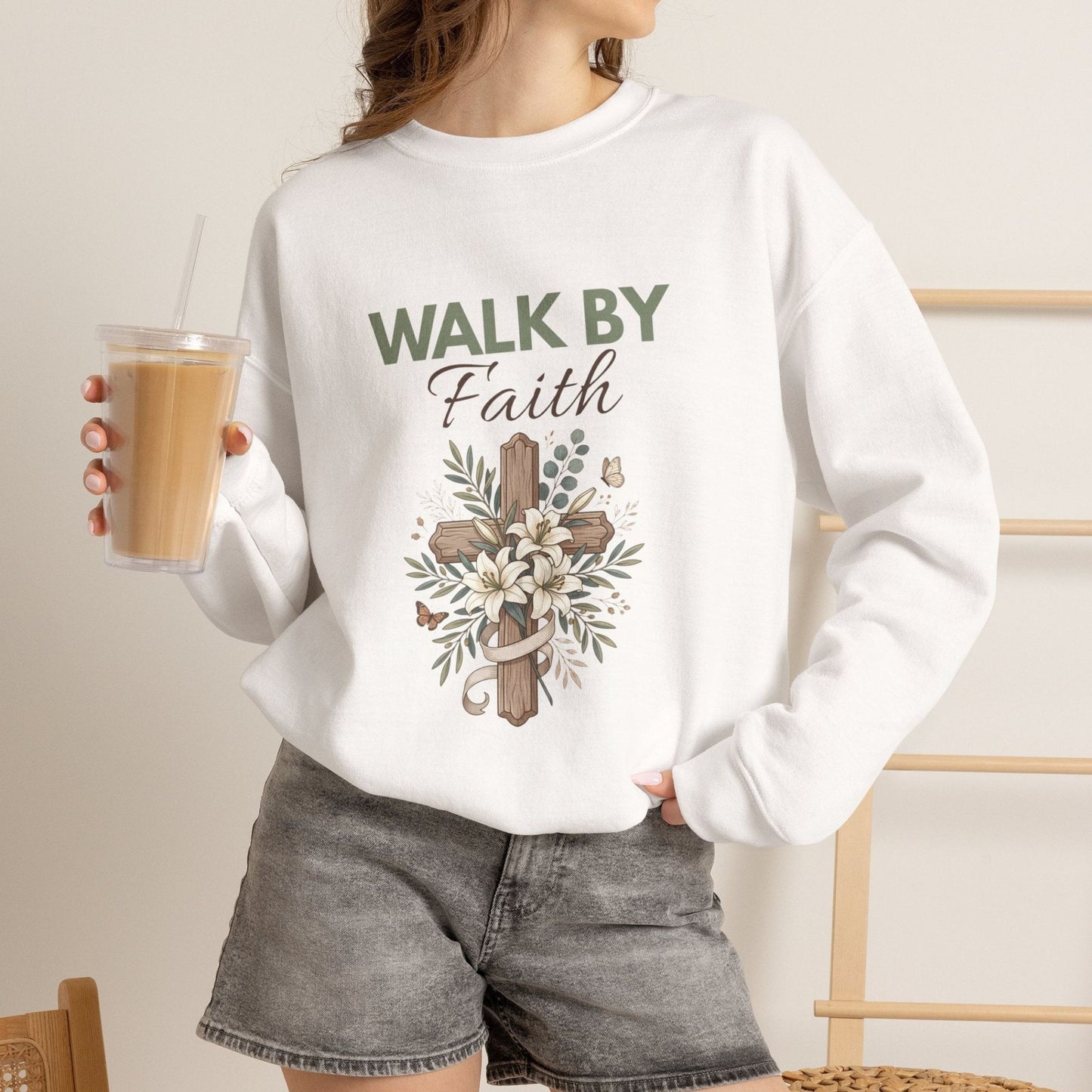 A woman is holding an iced coffee while wearing a white 'WALK BY Faith" crewneck sweatshirt.
