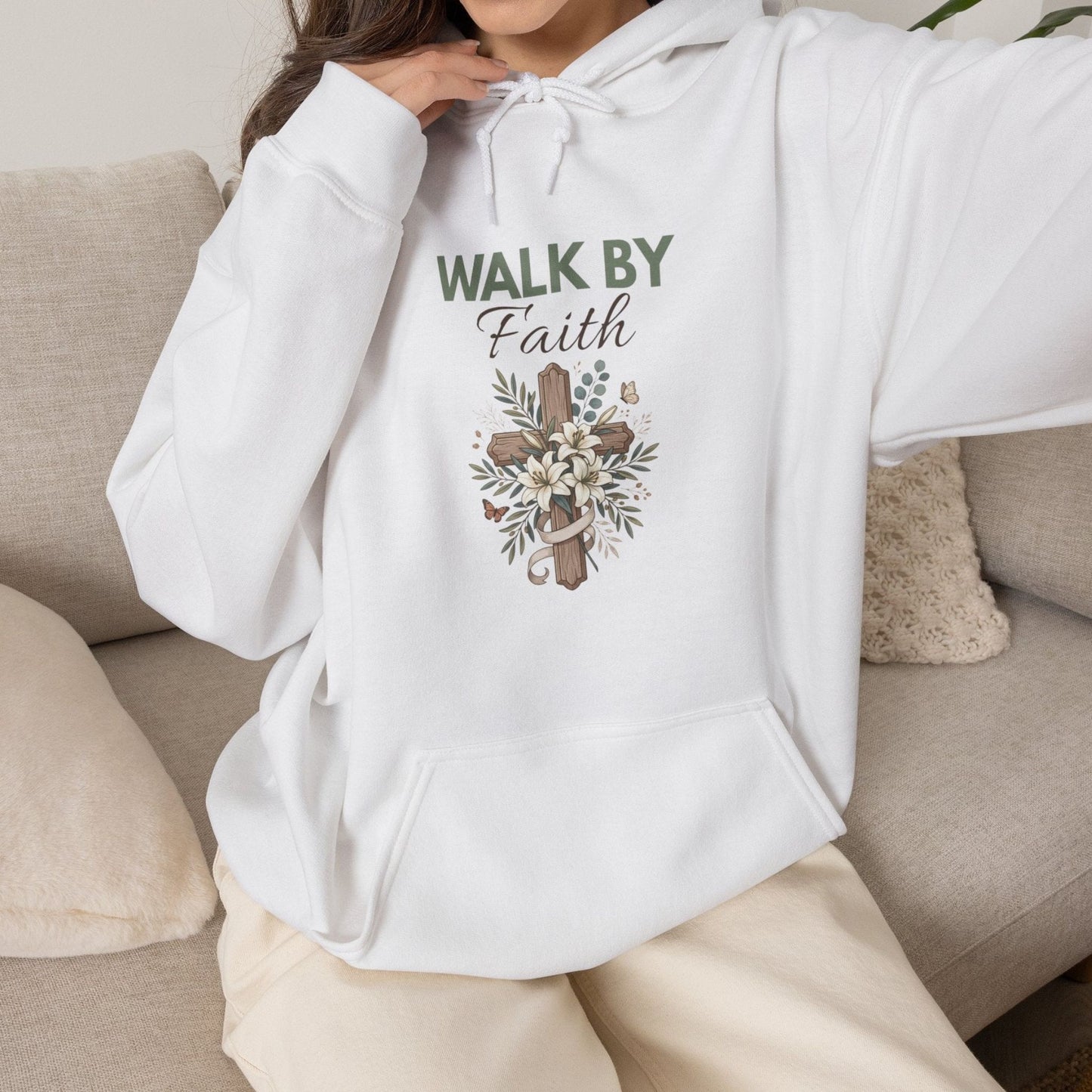 A woman sitting on a couch while wearing a white hooded sweatshirt with 'WALK BY Faith' on the front.