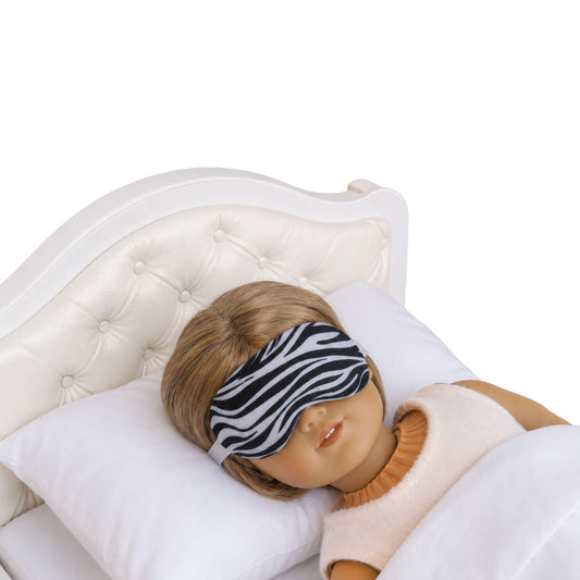 An 18-inch doll wearing a zebra print doll sleep mask while lying on a white upholstered doll bed.