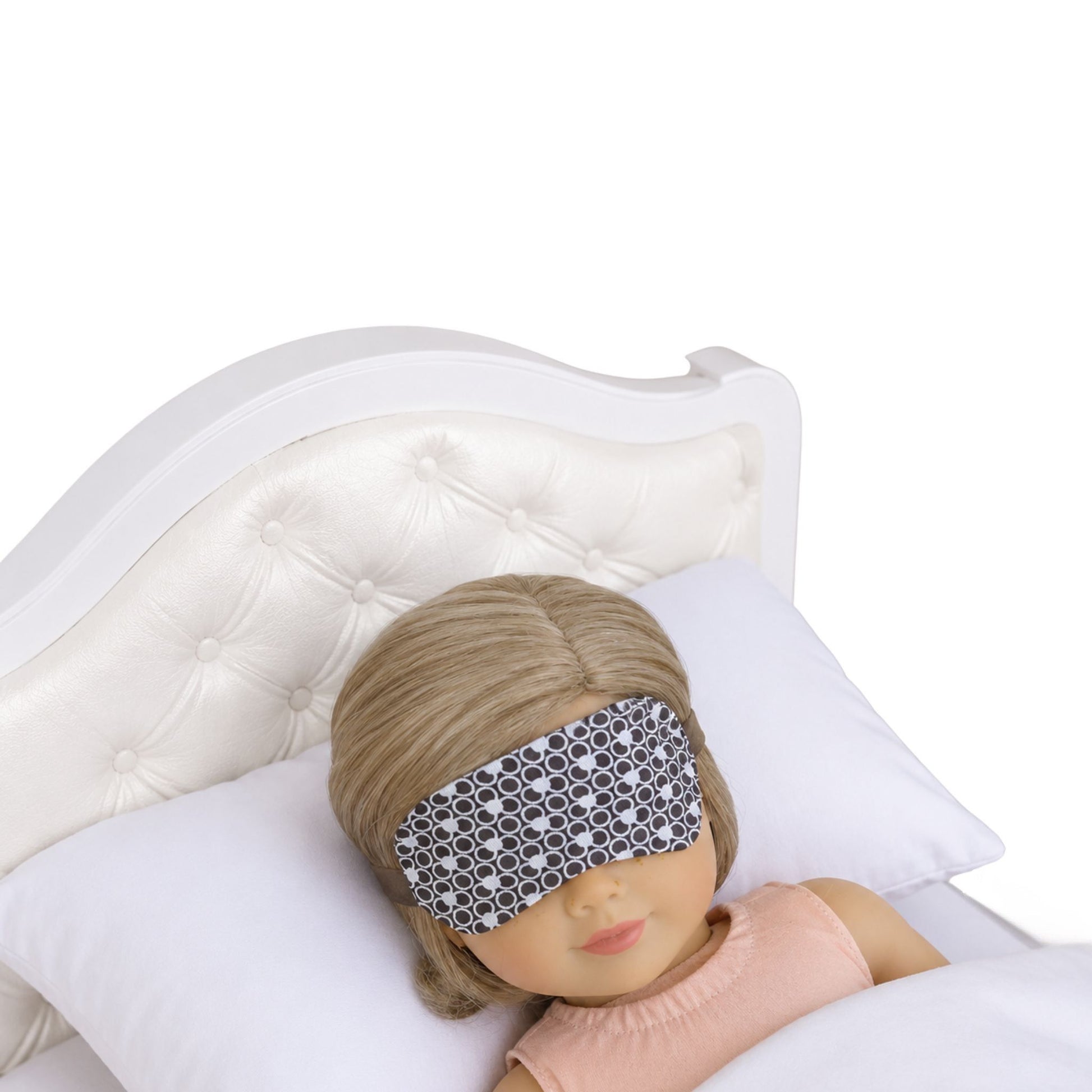 An 18-inch doll is wearing a white and brown print sleep mask while lying on a white doll bed.