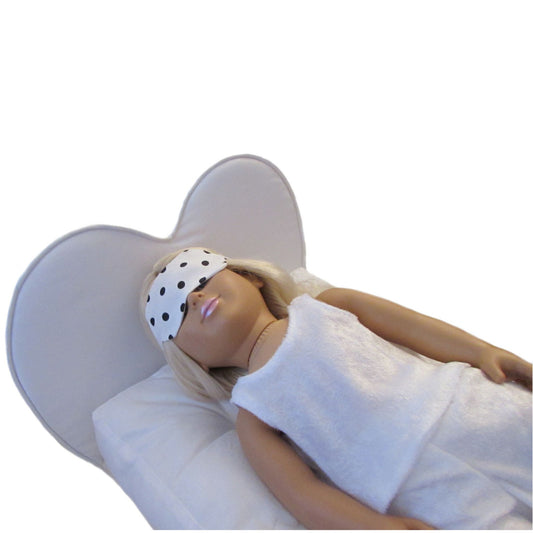 An 18-inch doll is wearing a sleep mask with black dots while lying on a white heart doll bed.