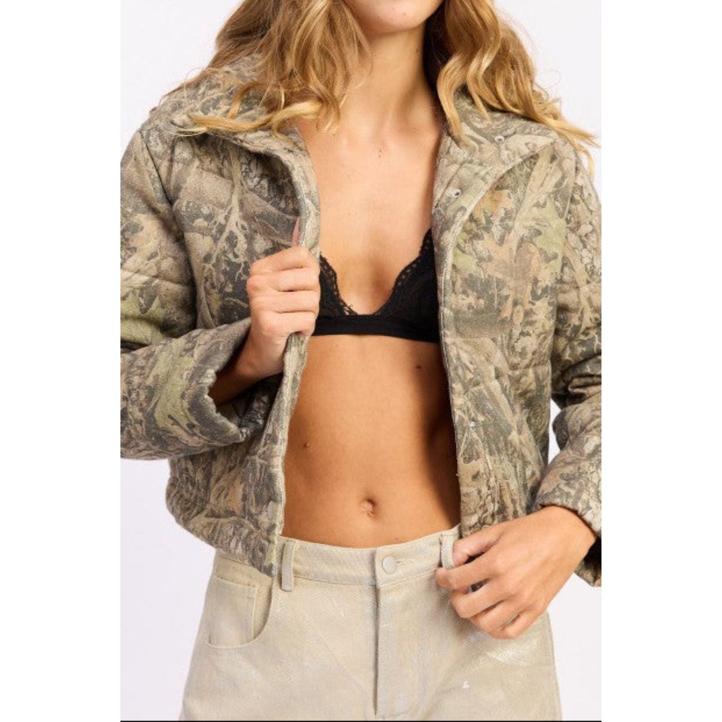 Person wearing a camouflage jacket over a black bra and beige pants on a white background
