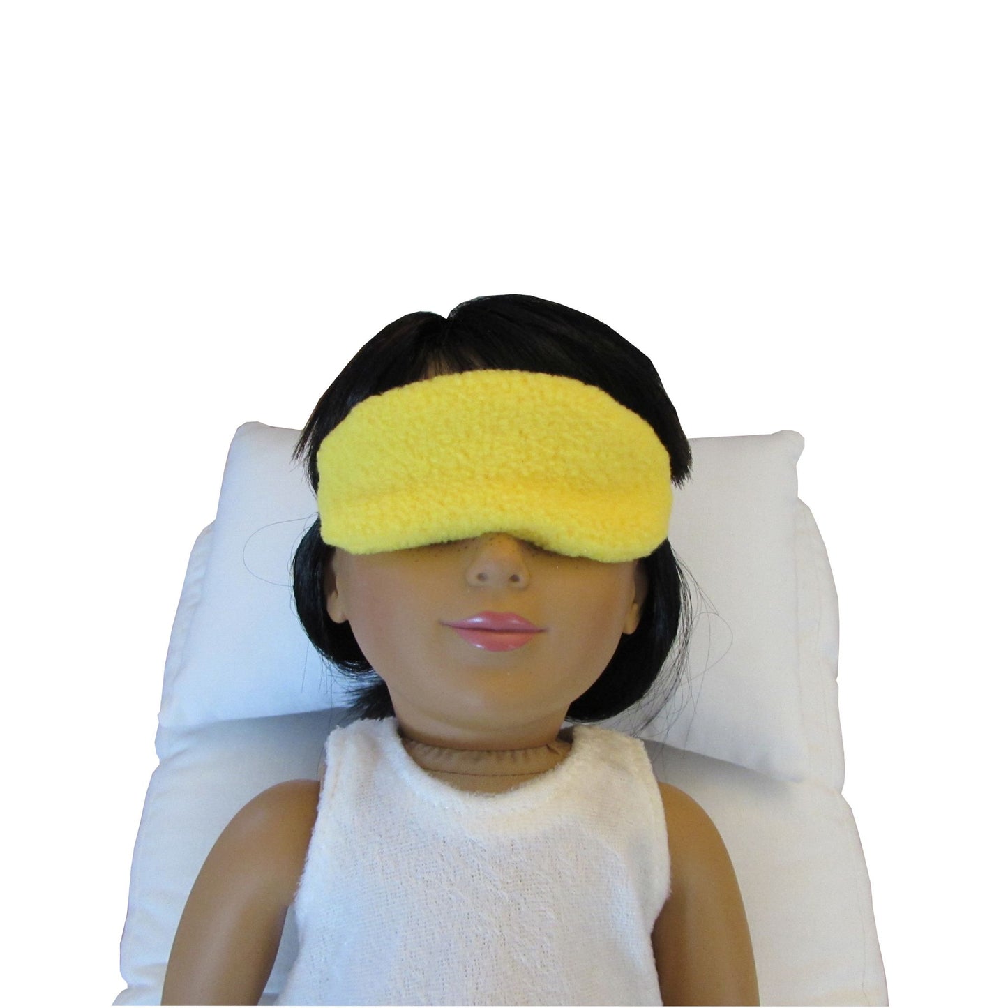 An 18-inch doll is wearing a yellow anti-pill fleece sleep mask while lying on a white doll pillow and a white doll mattress.