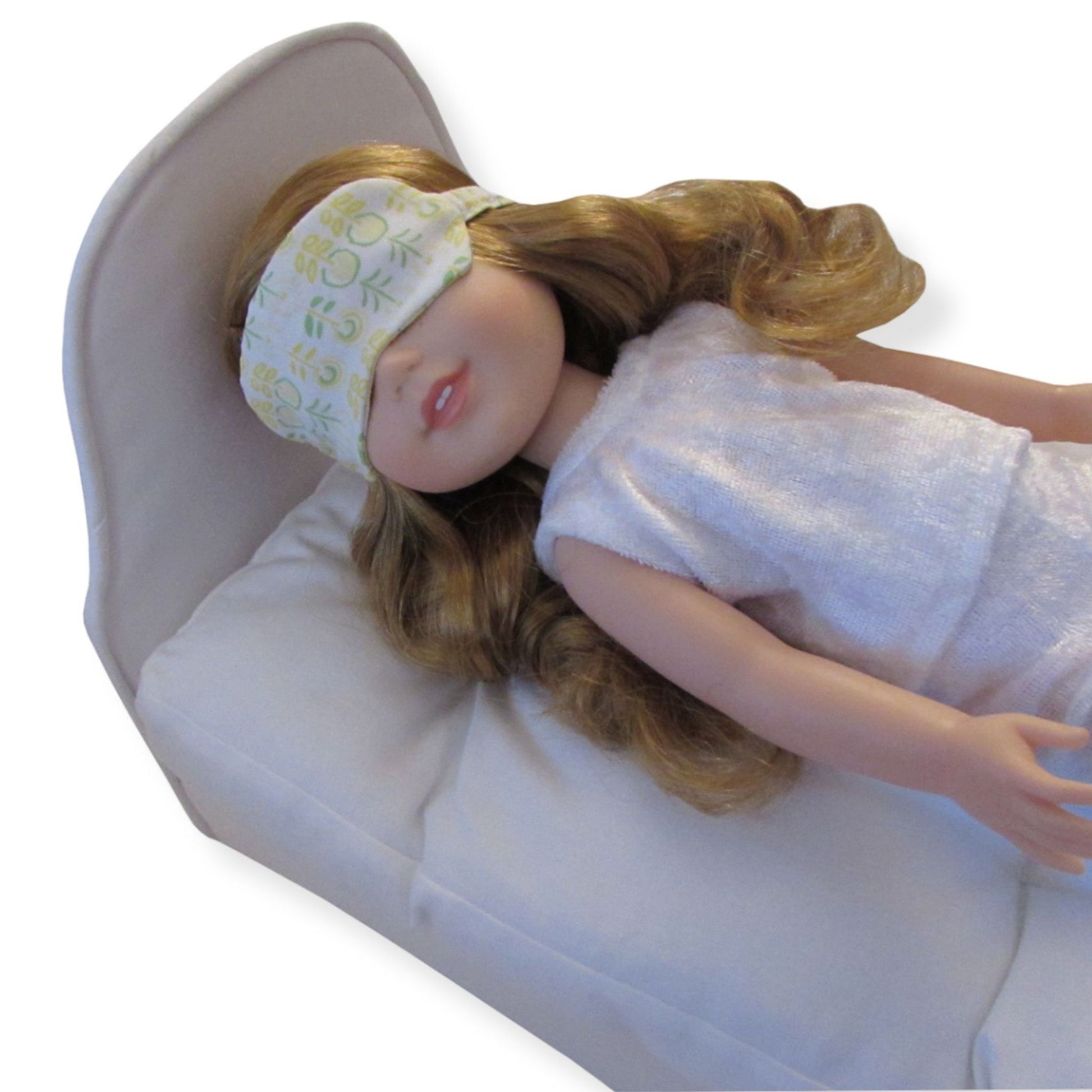 A 14.5-inch doll is wearing a yellow floral sleep mask while lying on a white doll bed.