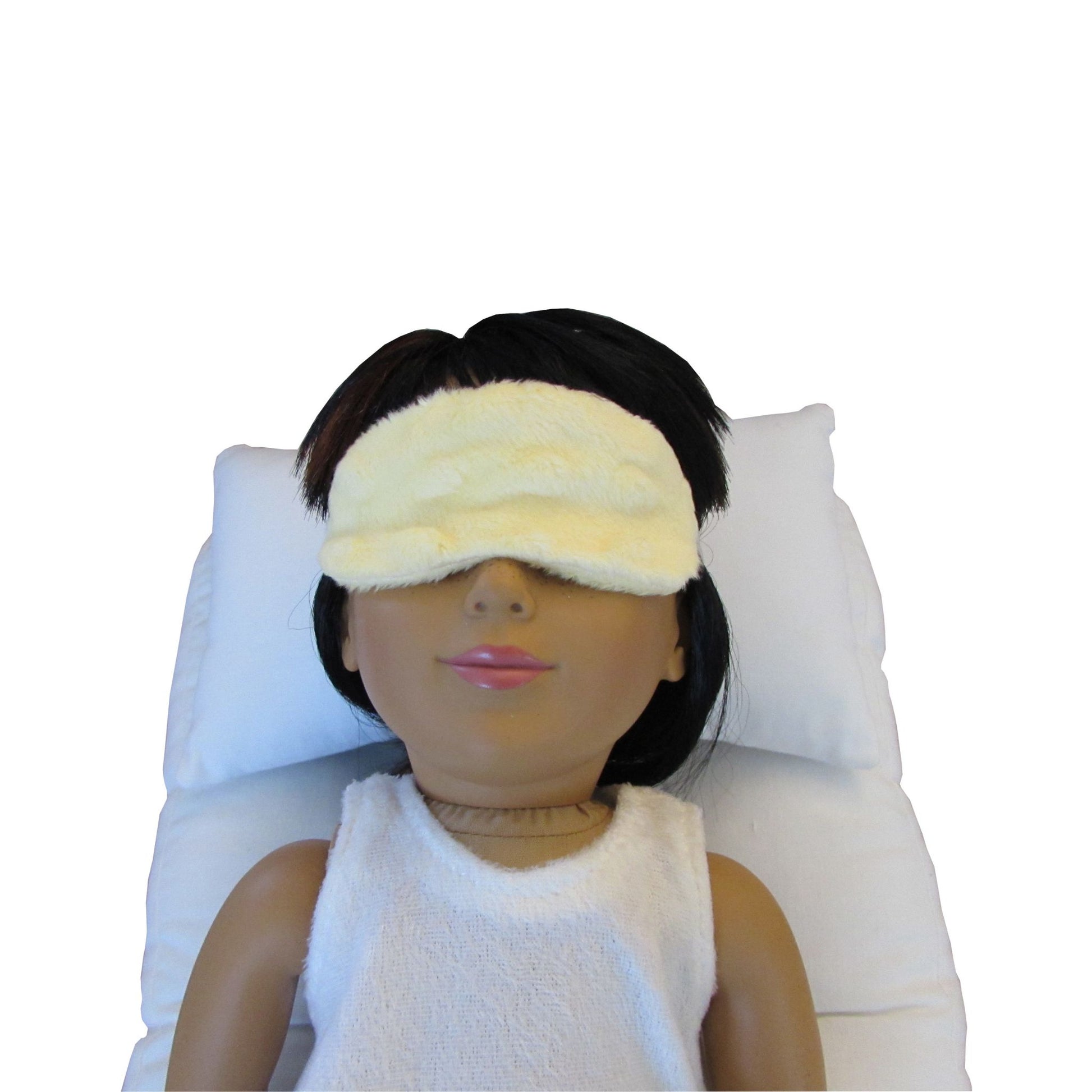 An 18-inch doll is wearing a yellow minky sleep mask while lying on a white doll pillow and a white doll mattress.