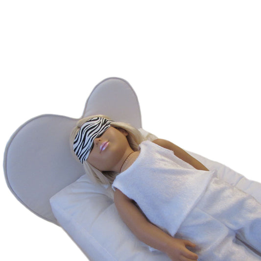 An 18-inch doll wearing a zebra print doll sleep mask and white pajamas while lying on a heart doll bed.