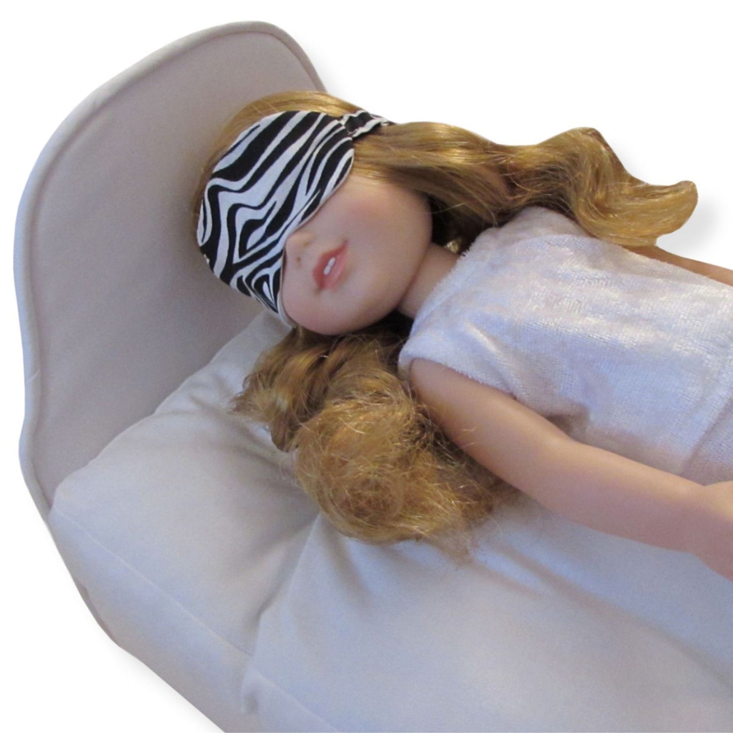 Doll with zebra-striped sleep mask lying on a white doll bed.